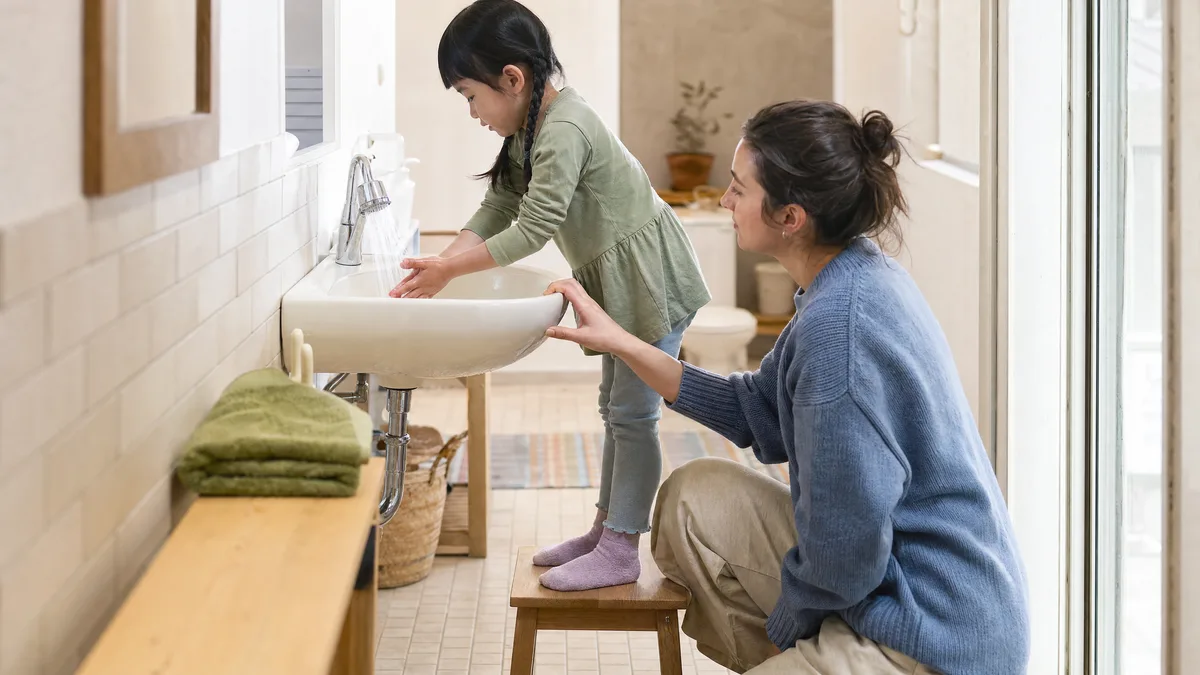 A girl standing on a step stool washes her hands while a therapist crouches to her level.