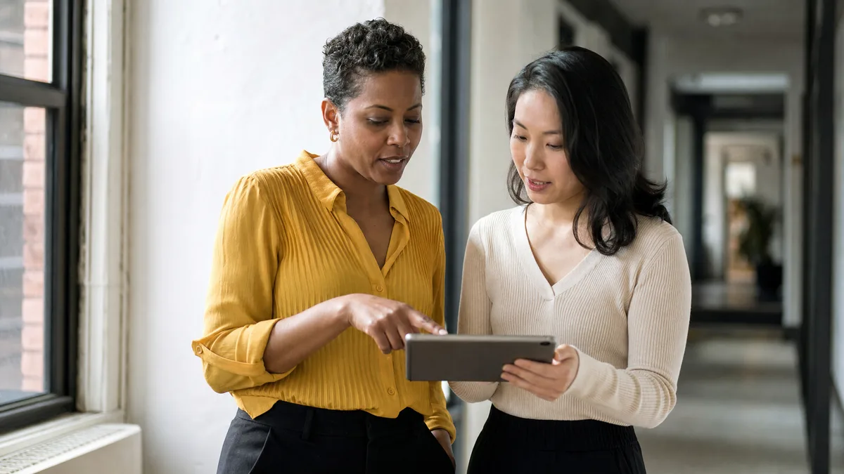 Two colleagues in a bright hallway, one showing the other something on a tablet.