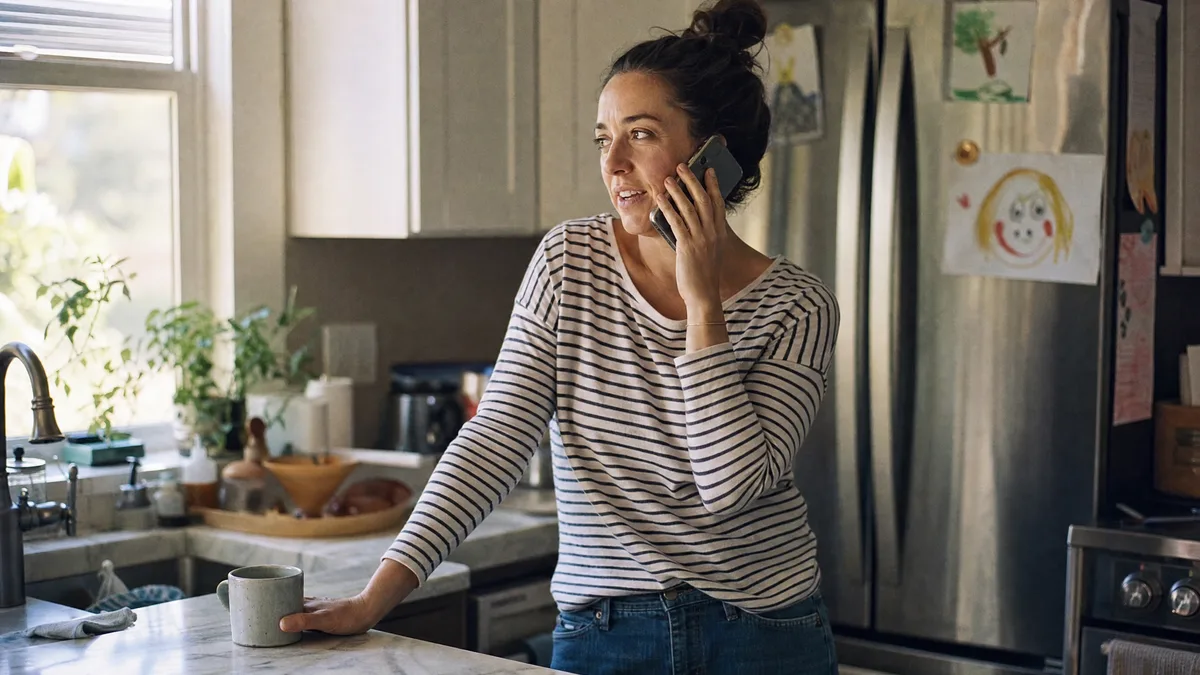 A parent standing in their kitchen on the phone, morning light coming through the window.